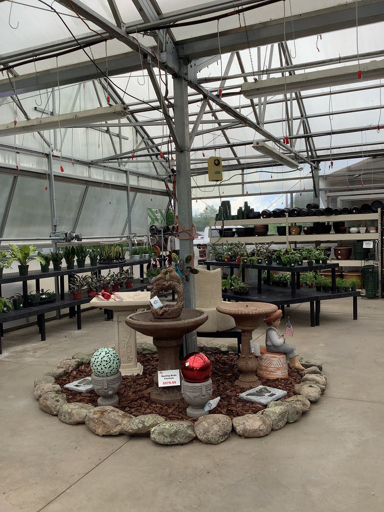 Foreground display of bird baths, garden spheres, and decorative statues. Background features plants and plant pots.